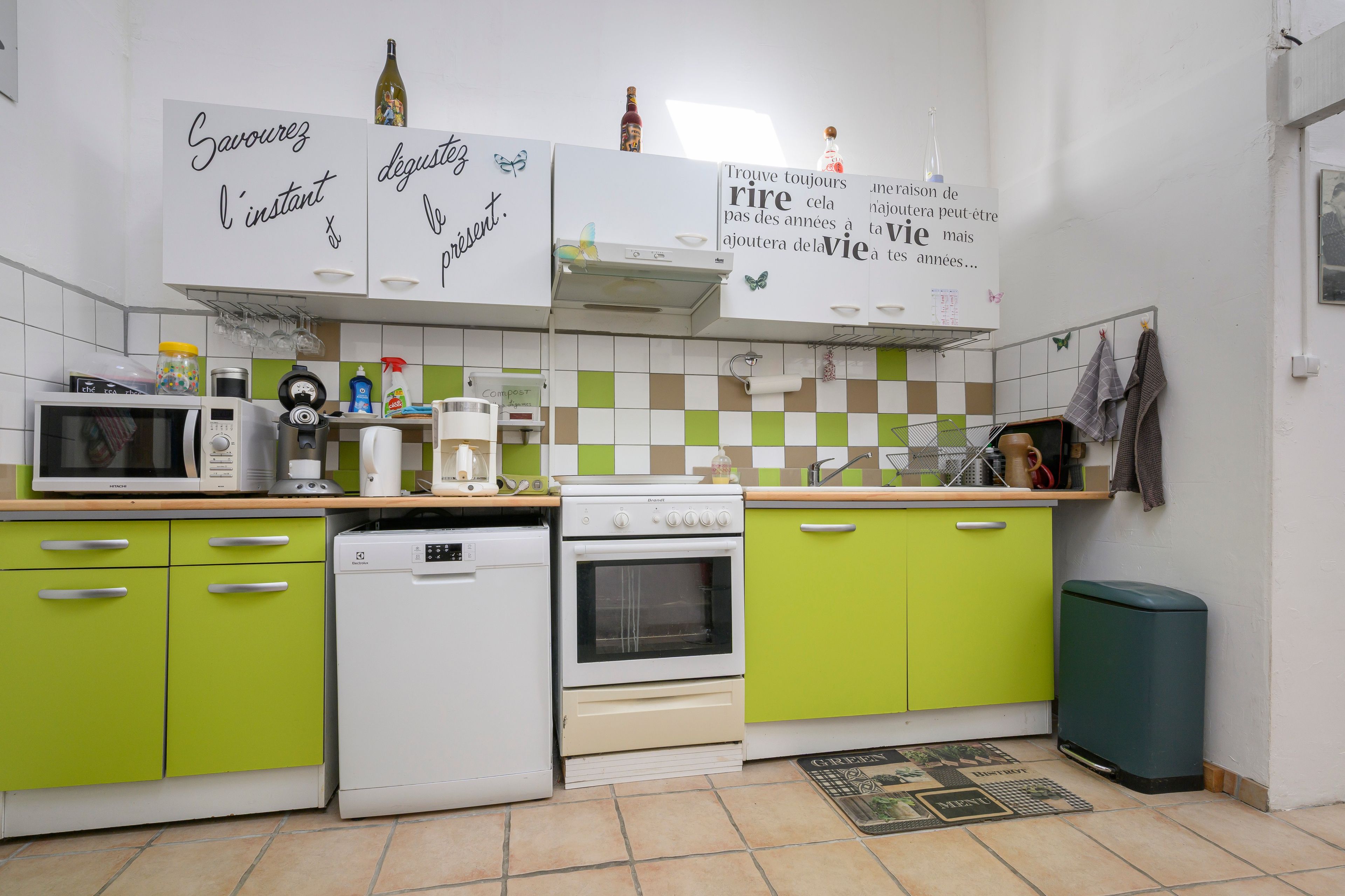 A kitchen with a microwave, coffee maker, and a stove. There are green cabinets and a trash can.