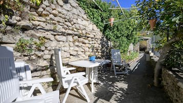 A stone wall with a garden area featuring white plastic chairs and a table, a narrow pathway, and a view of a mountain in the distance.