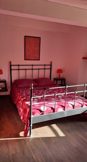 A bedroom with a metal bed frame, a red bedspread, two bedside tables with lamps, and a framed picture on the wall.