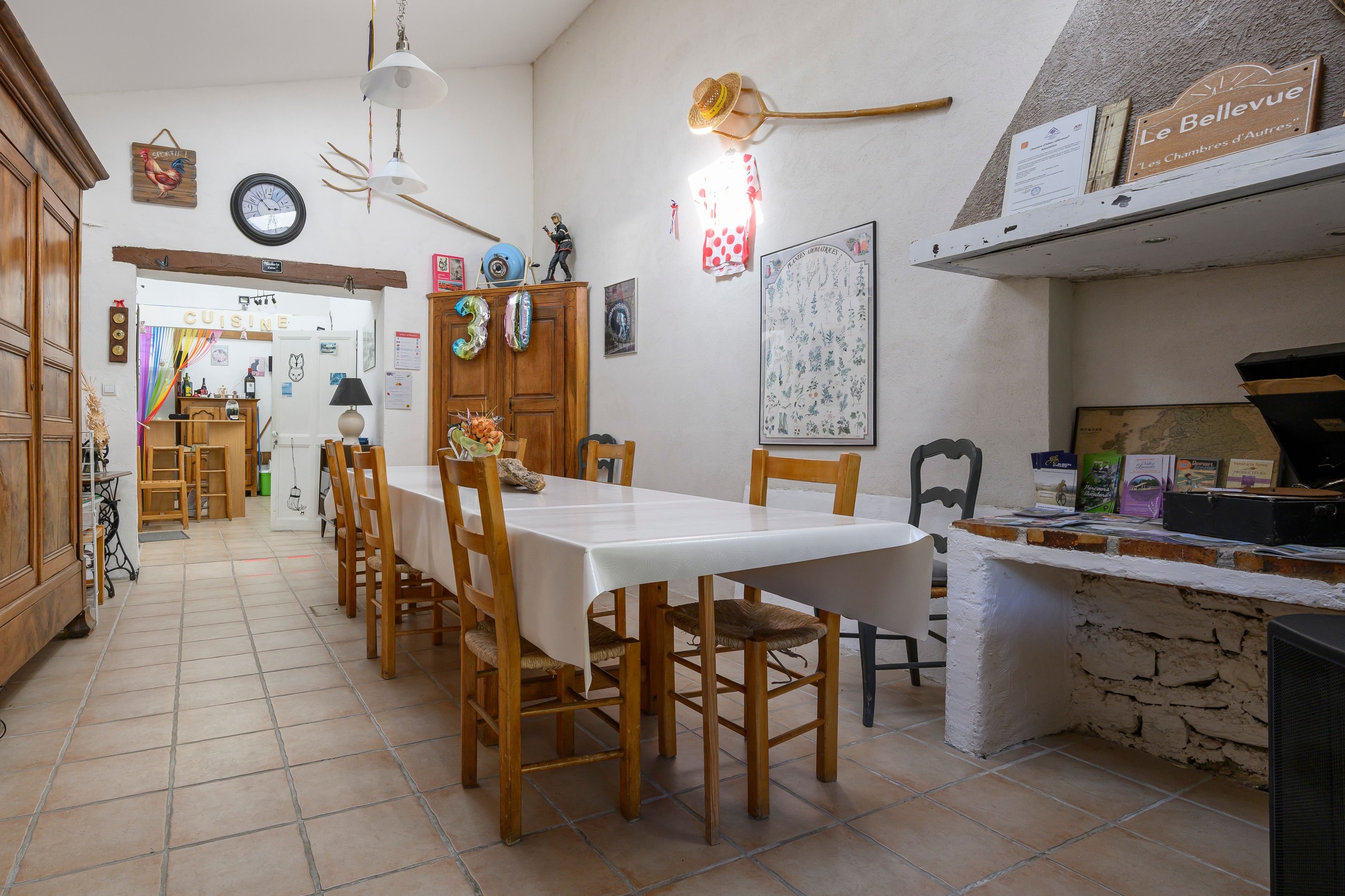 Un espace repas avec une longue table et des chaises, un coin cuisine avec un mur en pierre, et divers éléments de décoration, notamment un panneau représentant un coq et une horloge.