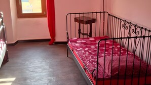 A bedroom with a metal bed frame, a wooden table, a window with a red curtain, and a framed picture on the wall.