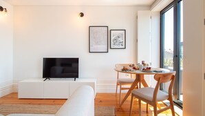 Una sala de estar moderna con un televisor de pantalla plana sobre un mueble blanco, una mesa de comedor con sillas y dos cuadros en la pared.
