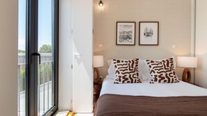 Una habitación de hotel con un ventanal grande, una cama con cubrecama marrón, dos almohadas de estampado de leopardo, dos lámparas de pared y dos cuadros en la pared.