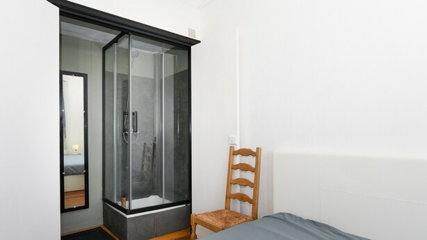 A bedroom with a bed, a wooden chair, a glass shower enclosure, and a mirror. - Spacious T2 with Unobstructed View (Lorient)