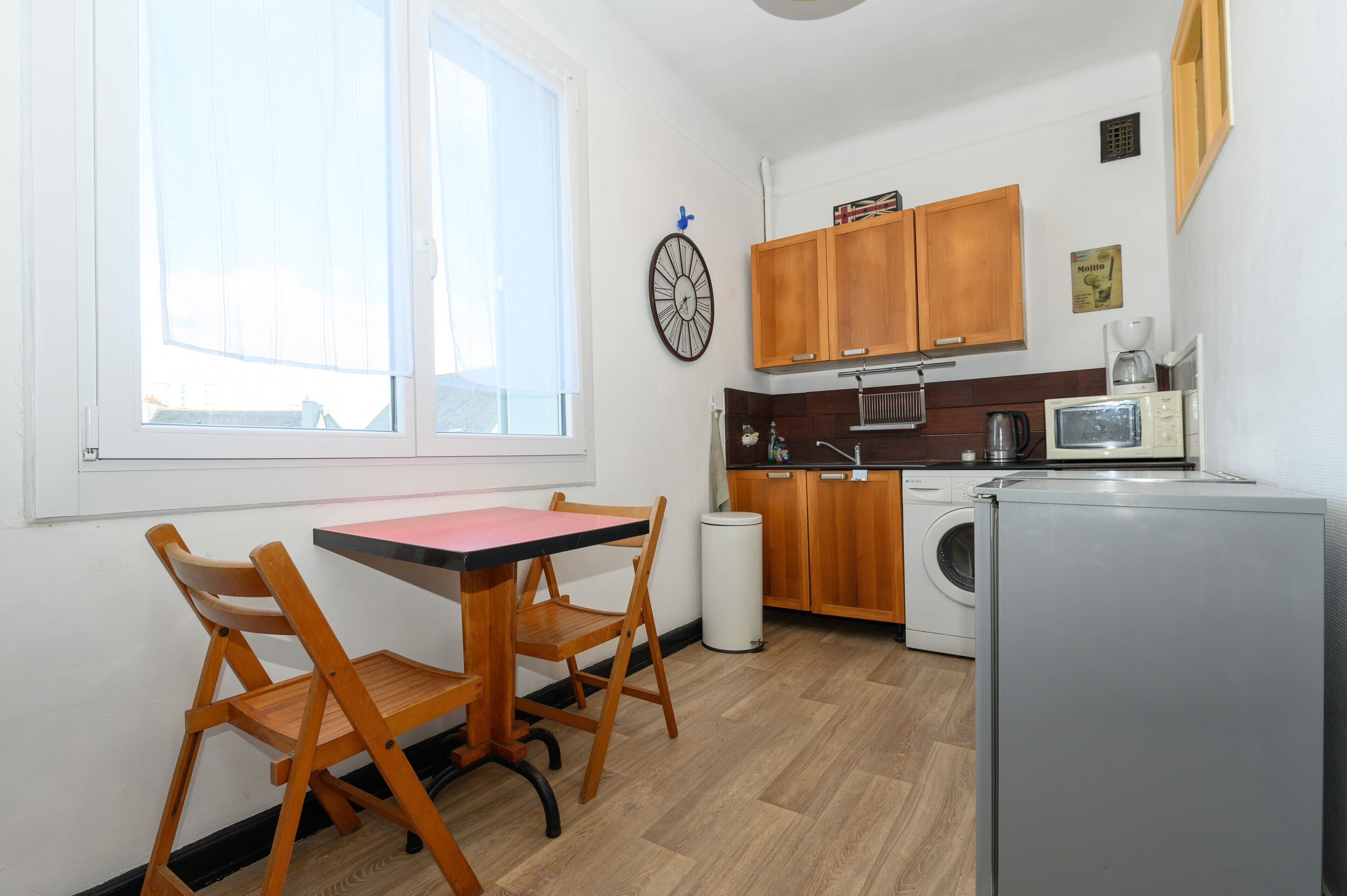 Een kleine keuken met houten kastjes, een wasmachine, een magnetron en een eethoek met een tafel en stoelen.