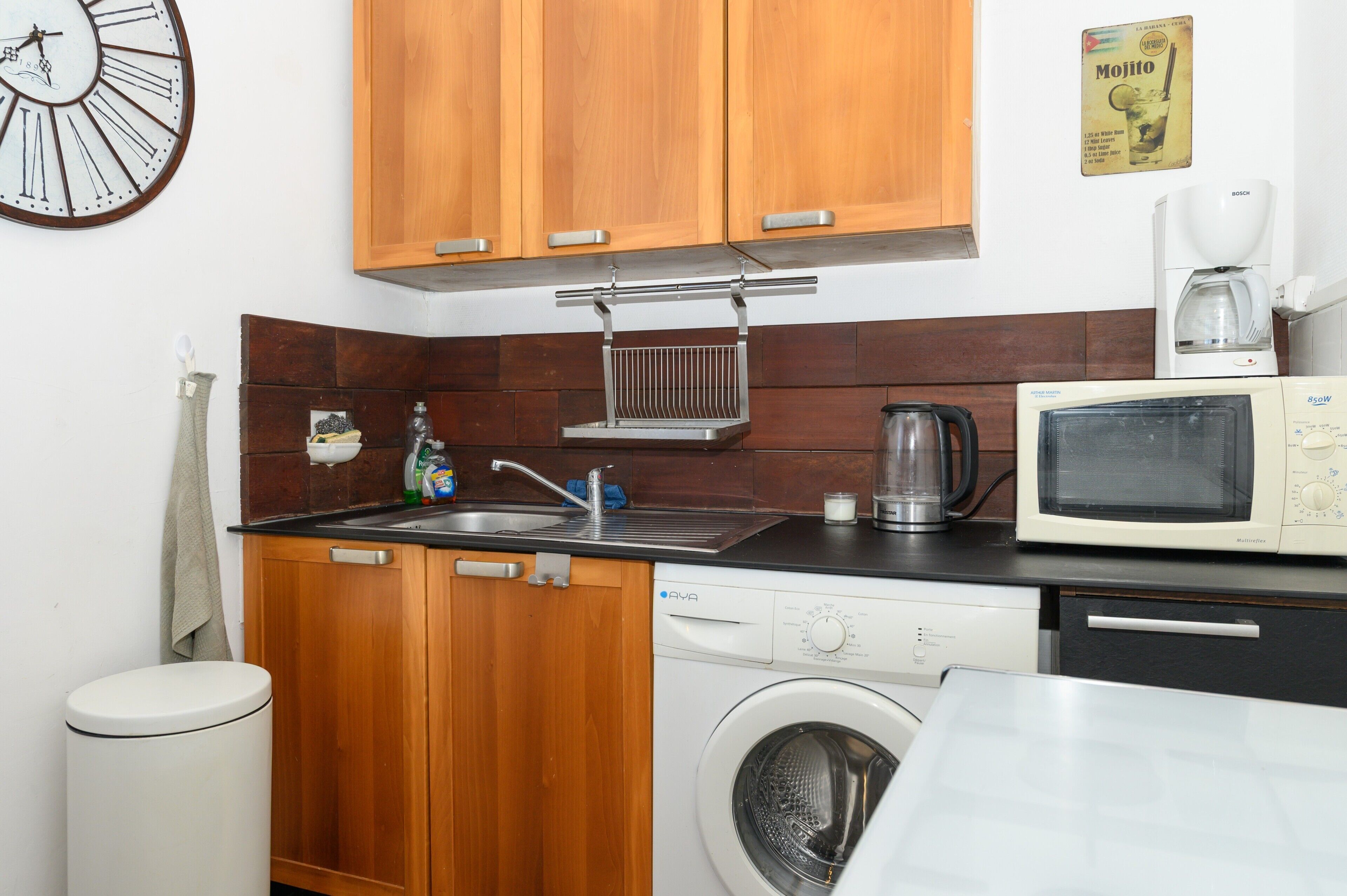 A compact kitchen with wooden cabinets, a washing machine, a microwave, and a coffee maker.