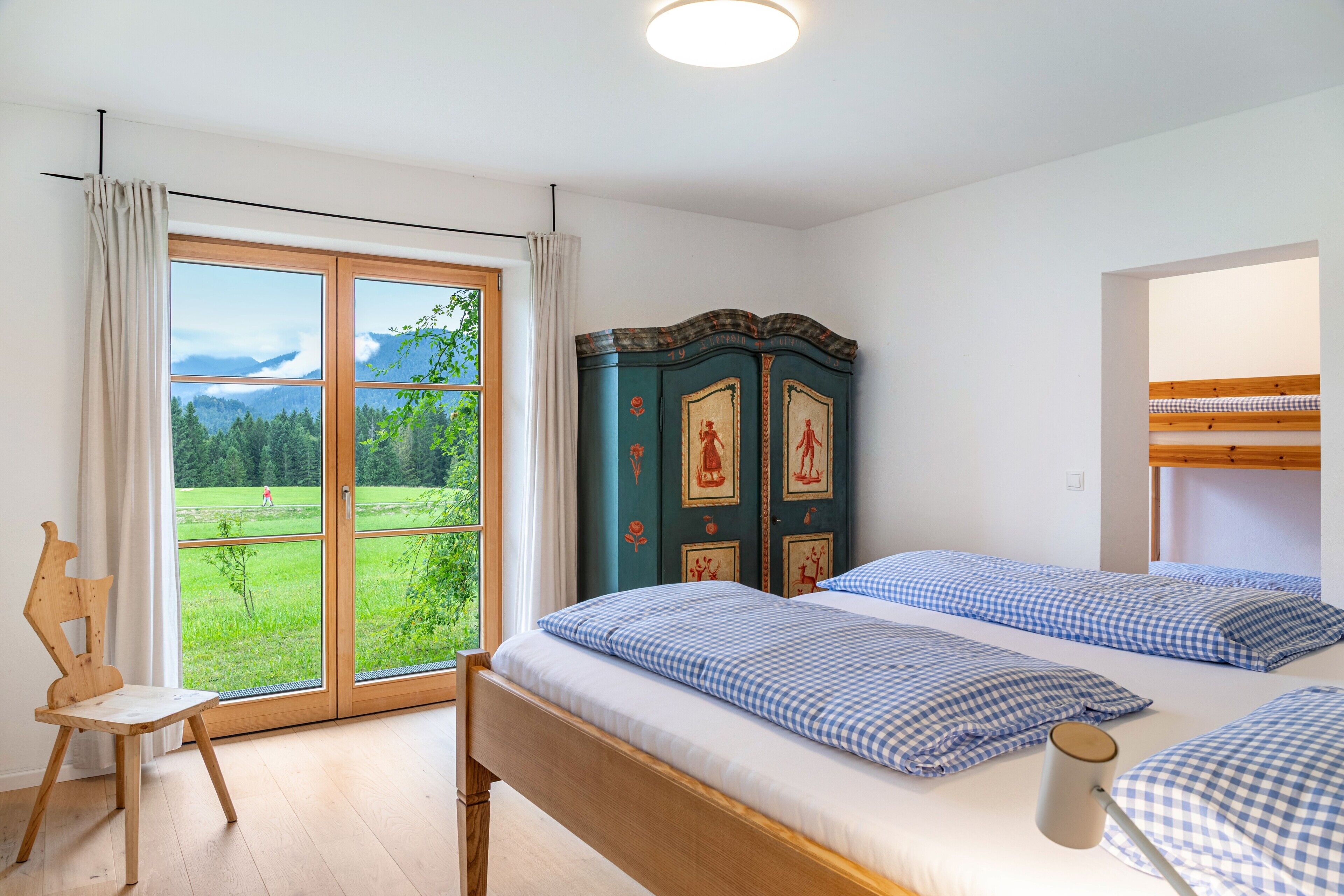 A bedroom with a large window, a wooden chair, a bed with checkered bedding, and a decorative cabinet.