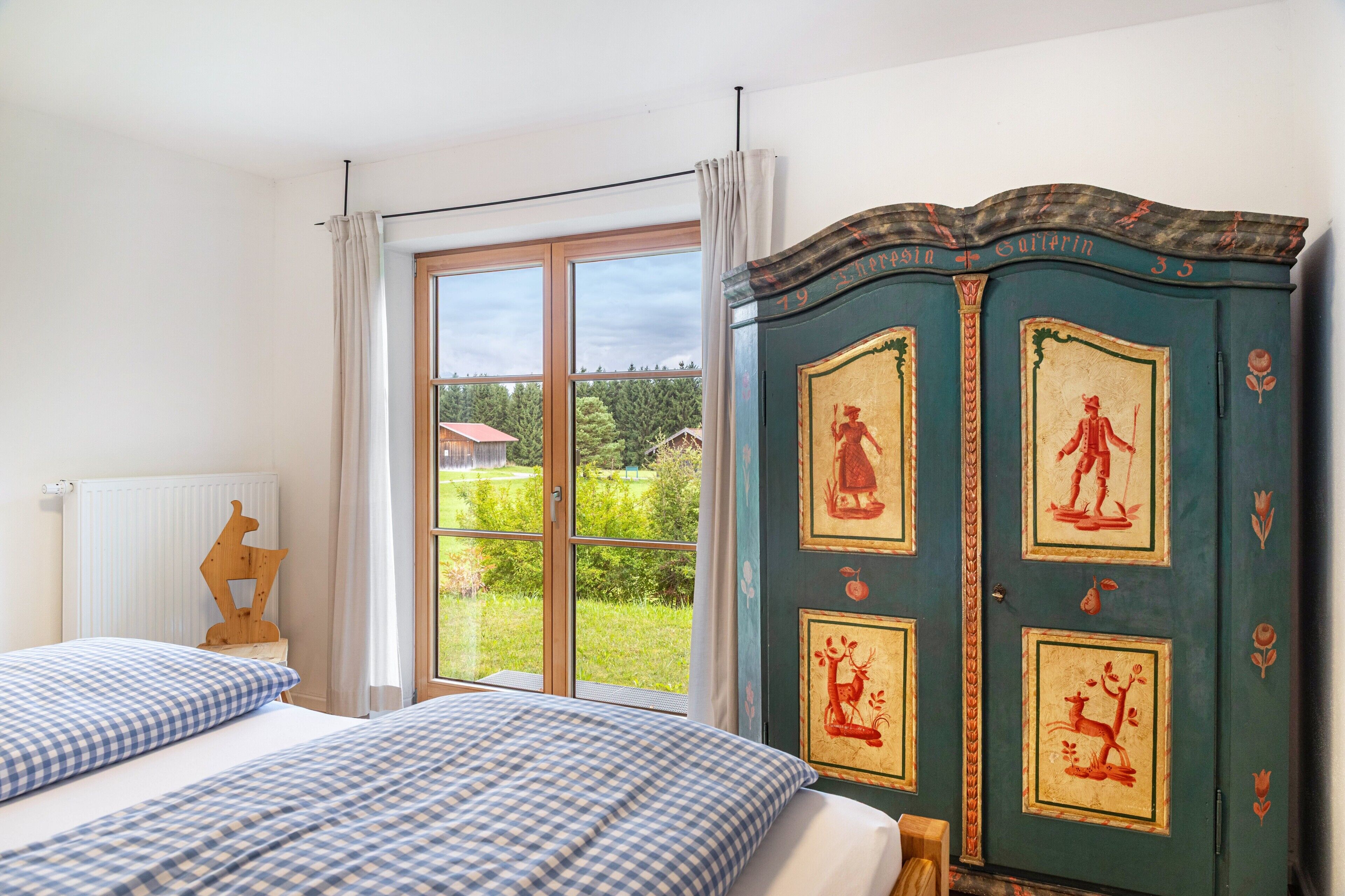 A bedroom with a large window, a bed with checkered bedding, and a green cabinet with artwork.