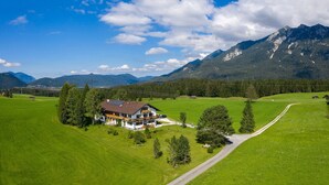 A large house with a sloped roof, surrounded by green fields and mountains. - Apartment '180 Grad' with Mountain View, Private Terrace and Wi-Fi (Mittenwald)