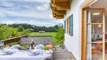 Ein Essbereich im Freien mit Tisch für Zwei, einer Holzbank und Blick auf eine grüne Landschaft mit Bäumen und einem Haus in der Ferne.