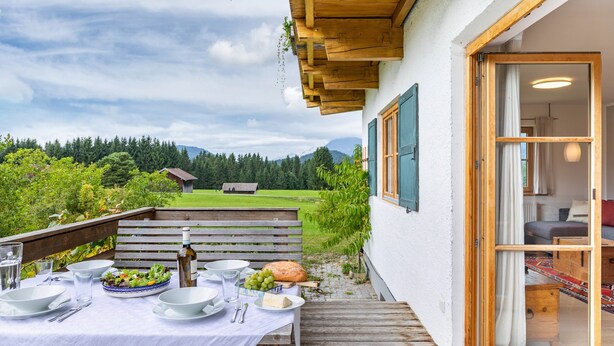 An outdoor dining area with a table set for two, a wooden bench, and a view of a green landscape with trees and a house in the distance. - Apartment '180 Grad' with Mountain View, Private Terrace and Wi-Fi (Mittenwald)