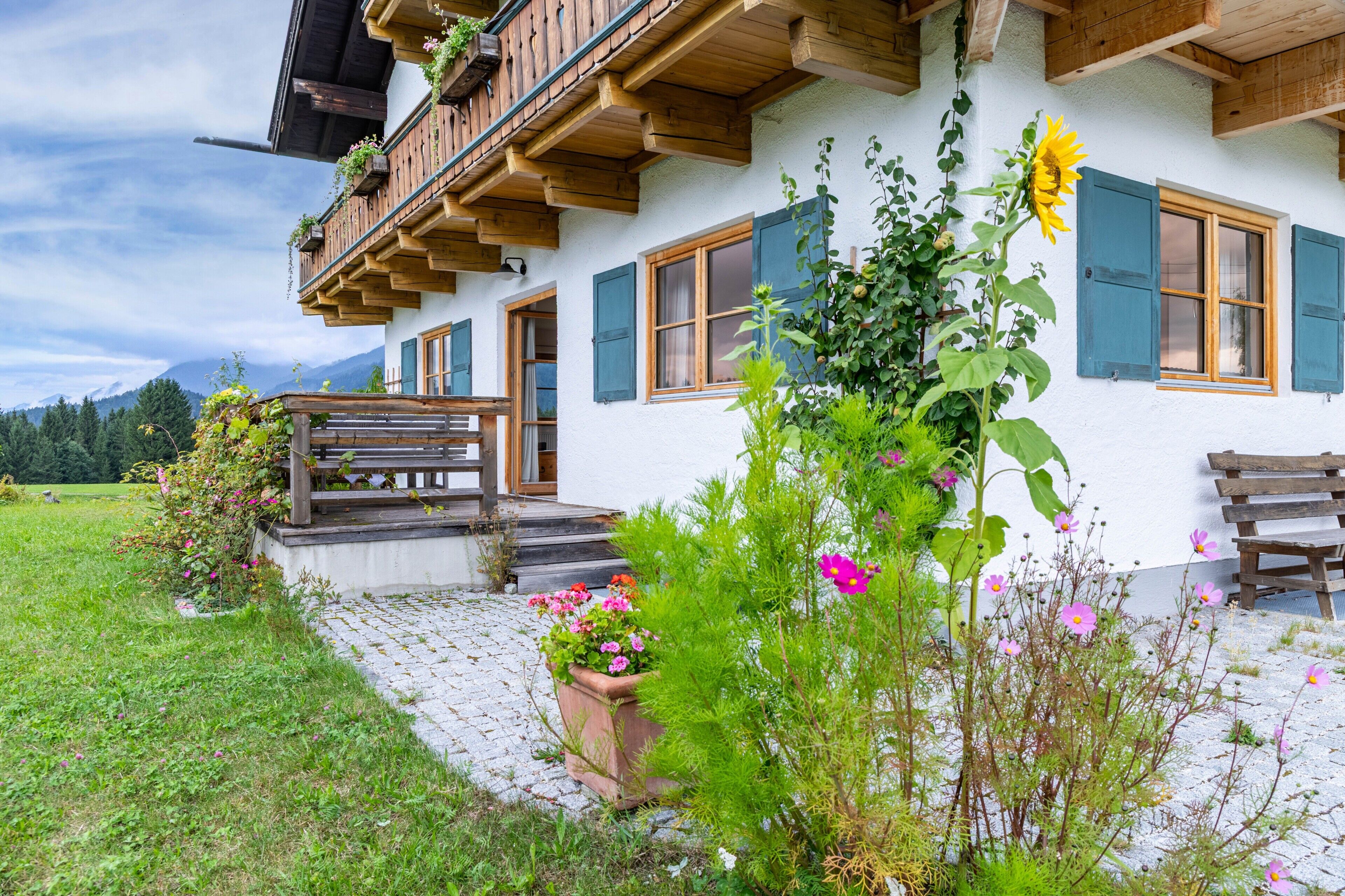 En traditionell chalet med träbalkonger, en entré med stenläggning och en trädgård med blommor och en solros.