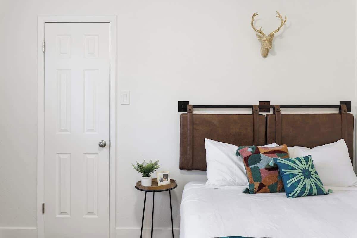 A bedroom with a white door, a bed with a brown headboard, and a mounted deer head on the wall.