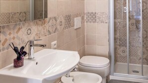 A bathroom with a white sink, two toilets, a glass shower enclosure, and patterned tiles. - B&B Stern (Porto San Giorgio)