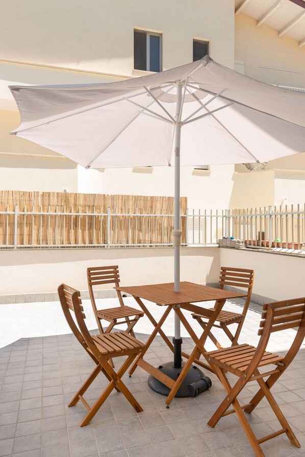 A patio with a round wooden table and four chairs, an umbrella, and a bamboo fence in the background. - B&B Stern (Porto San Giorgio)