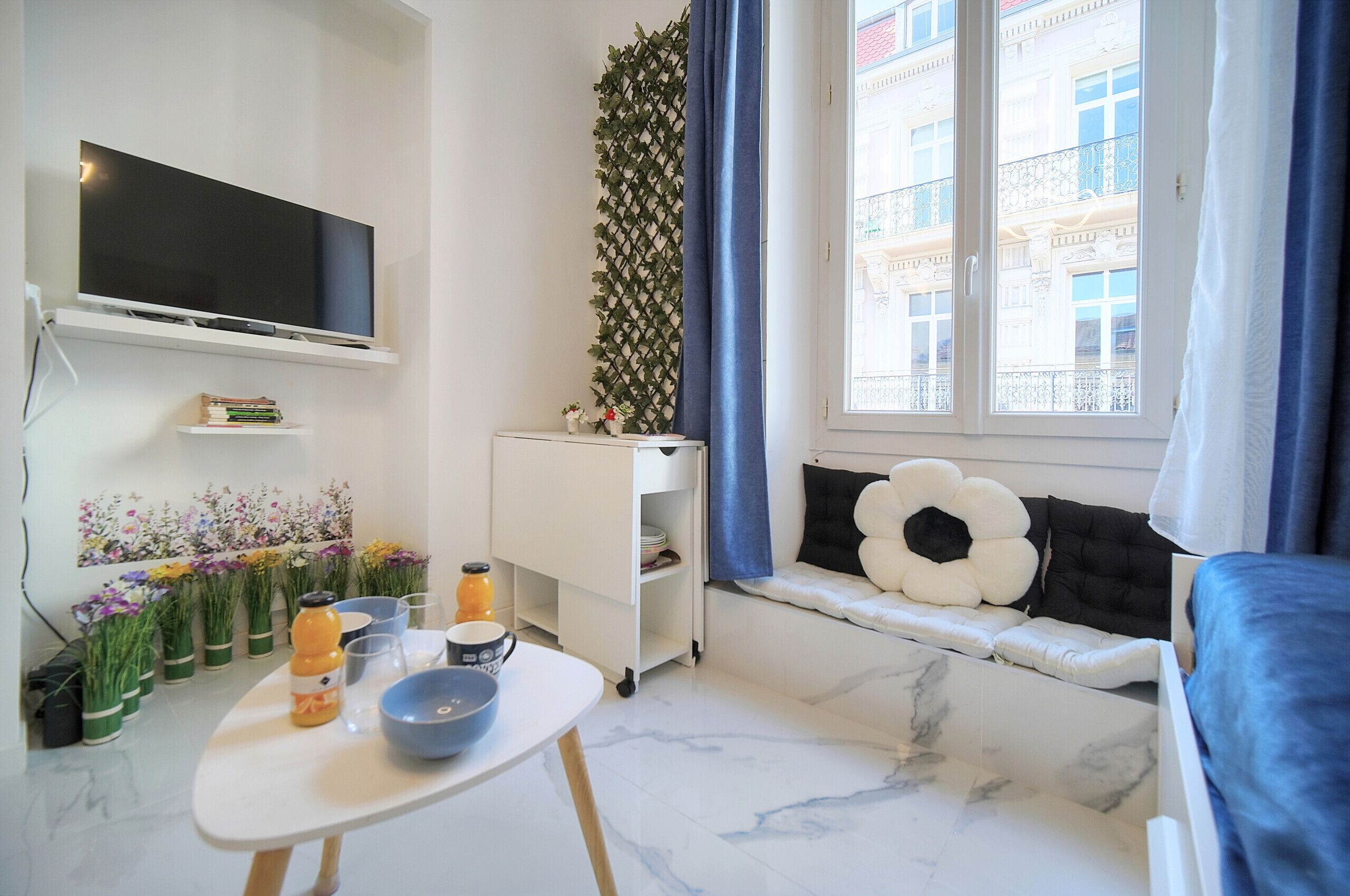 A modern living room with a flat-screen TV, a small table with a bowl and a bottle, a sofa with a decorative pillow, a window with curtains, and a plant wall.