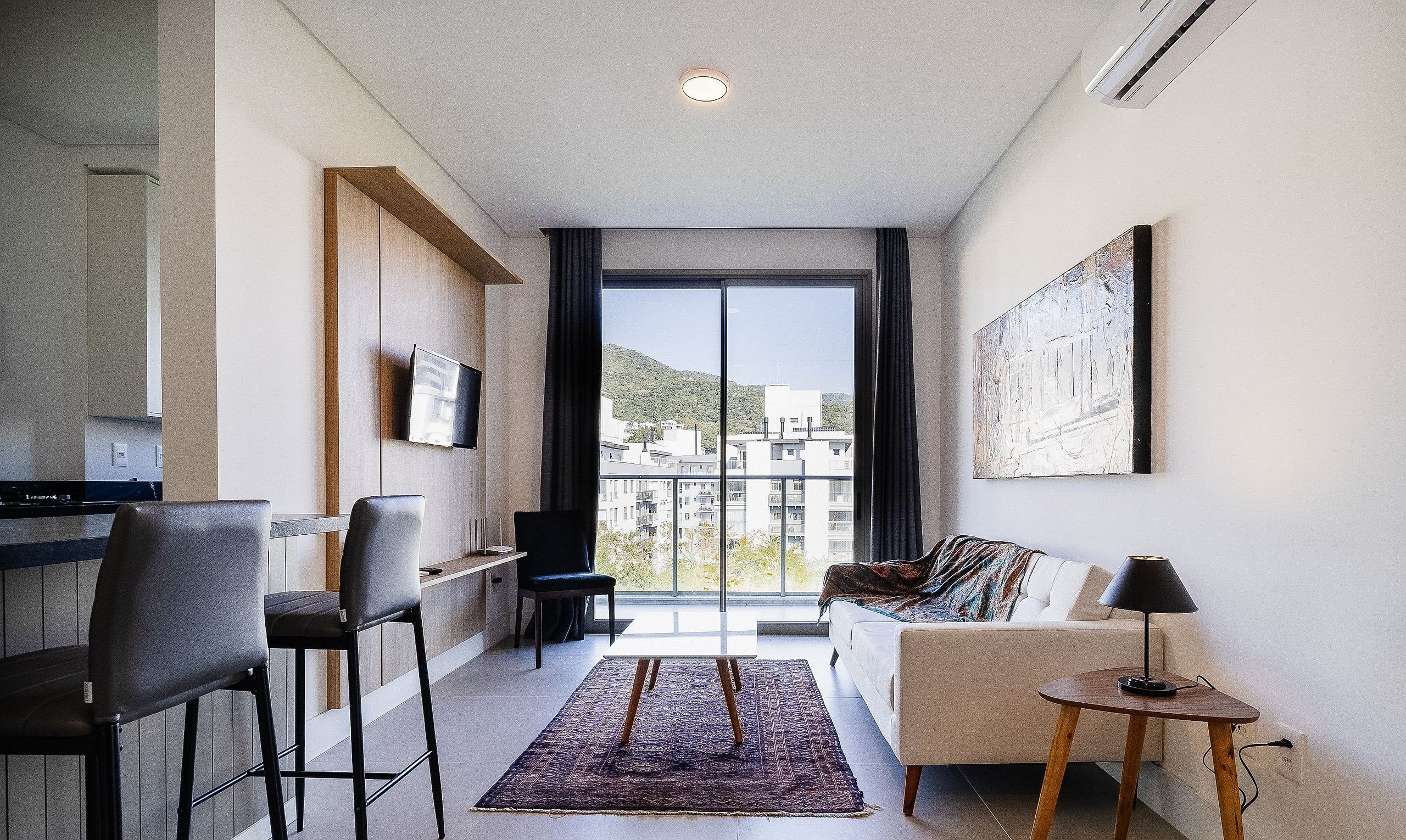 A modern living room with a sofa, chairs, a TV, and a view of buildings through a large window.
