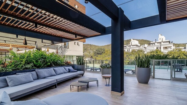 A modern outdoor seating area with a sofa and tables, surrounded by potted plants and overlooking a scenic view of buildings and hills. - Stay in a Building With a Pool, Gym, and Leisure - SSL (Florianópolis)