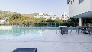 A swimming pool with a patio area, surrounded by a metal fence and trees.