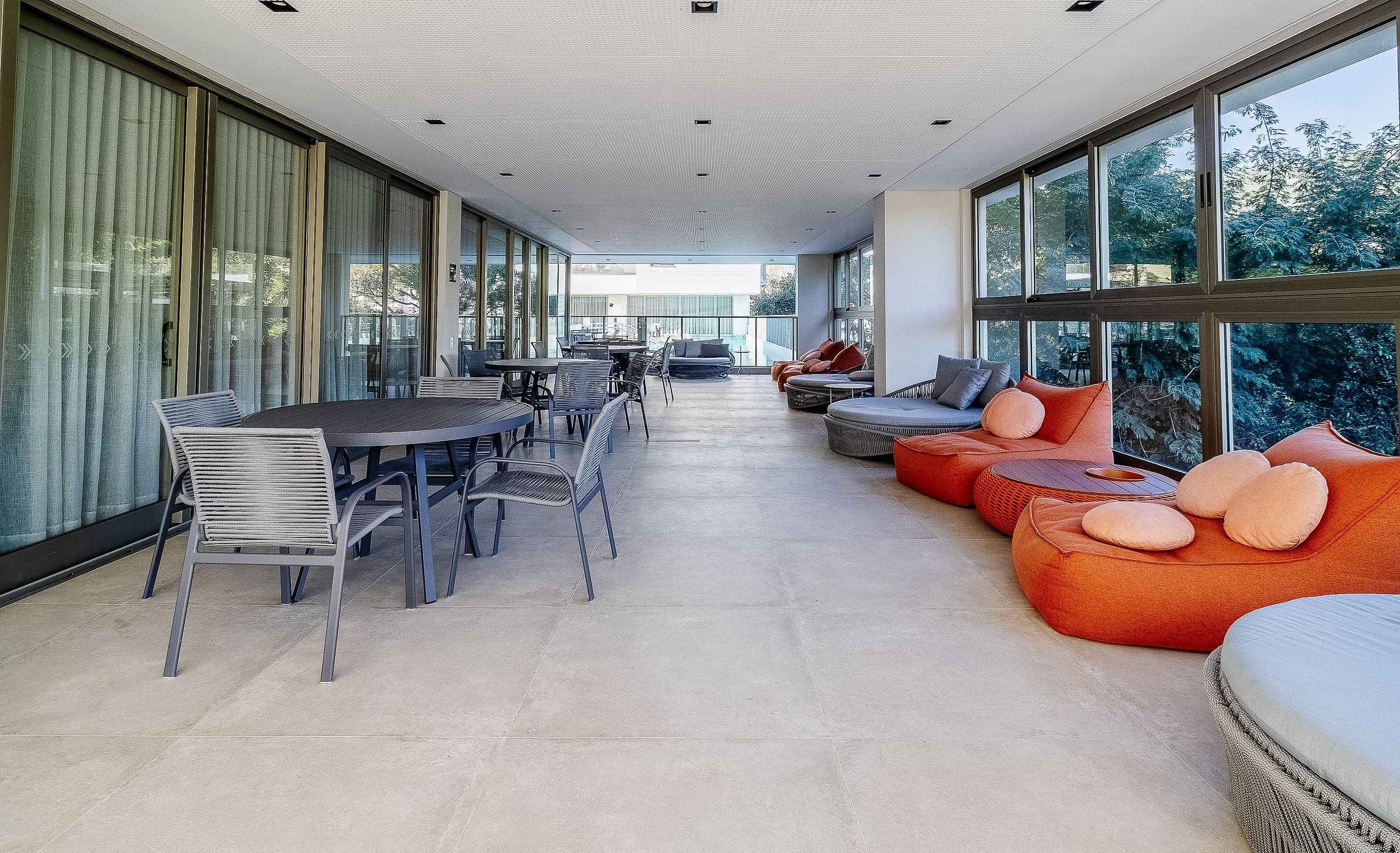 A spacious indoor area with a long table and chairs, a sofa with cushions, and large windows offering a view of greenery outside.