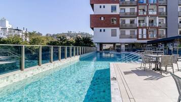 Una piscina con agua azul cristalina, rodeada por una barandilla de vidrio y un patio con mobiliario exterior.