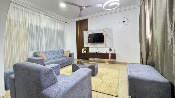 Una sala de estar moderna con un sofá gris, un televisor de pantalla plana montado en una pared de mármol, un ventilador de techo y un amplio ventanal con cortinas de gasa.