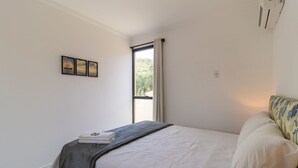 A bedroom with a bed, a window with curtains, a bedside table with a towel, and a wall-mounted air conditioner. - Apt With Barbecue in Lagoa da Conceicao Sro0002 (Florianópolis)