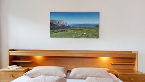 Un dormitorio con cabecera de madera, dos almohadas, una cama bien hecha, una mesita de noche con un libro y una lámpara, y un cuadro de paisaje en la pared.