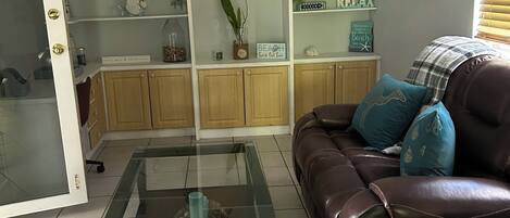 Sala de estar com mesa de centro de vidro, sofá de couro marrom com almofadas azuis e uma estante com objetos decorativos.