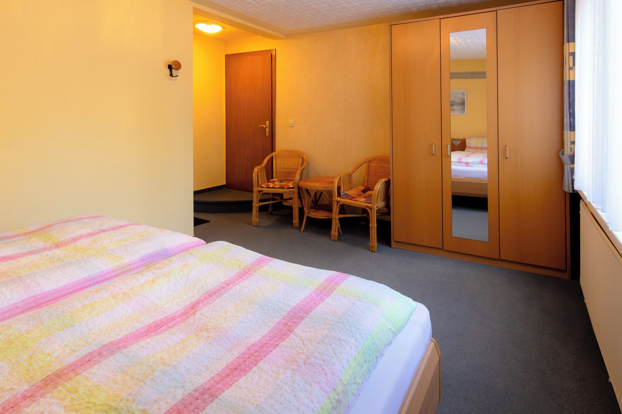 A hotel room with a bed, two wicker chairs, a wardrobe, and a door.