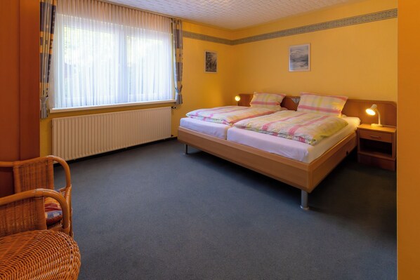 A bedroom with a double bed, a chair, a bedside table with a lamp, and a window with curtains.