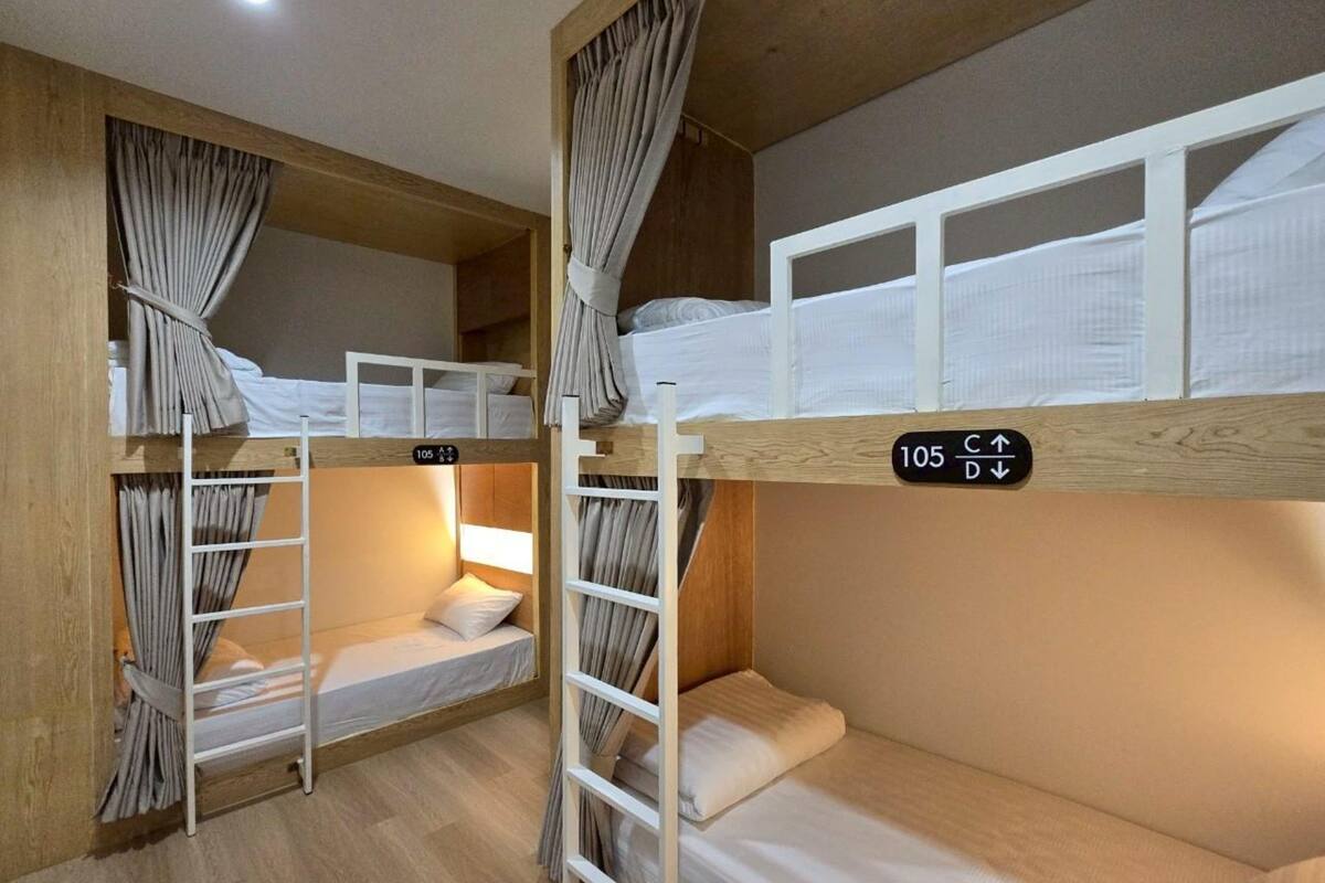 Quarto com beliches, paredes de madeira, escada e cortinas. Há placas com números de quarto e indicações de direção.