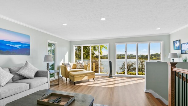 A spacious living room with a grey sofa, a beige armchair, and a wooden coffee table. Large windows offer a view of a lake and greenery. - Graceful Swan Retreat w/ Dock, Pool & Golf Access (Lake Junaluska)