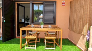 A wooden table with chairs and a potted plant in a sunny outdoor area with artificial grass. - Home2Book Charming Apt Poolside Retreat (Corralejo)