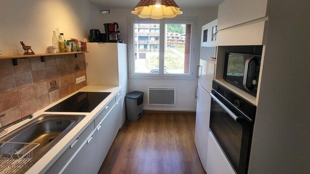 A compact kitchen with a built-in stove, microwave, and sink. It features a window with a view of the outside, a hanging pendant light, and a small trash bin.