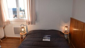 A bedroom with a dark quilted bed, a window with curtains, two bedside lamps, a radiator, and a wooden headboard.