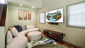 A modern living room with a sectional sofa, a flat-screen TV mounted on the wall, and a decorative wall panel. - Streamside | Nature-Lux w/ Hot Tub, Sauna, Firepit, Game Area, Beautiful Setting (Traverse City)