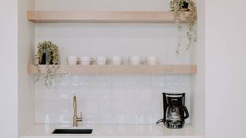 Cocina con armarios de madera, encimera blanca, cafetera negra y dos repisas con plantas y tazas blancas.