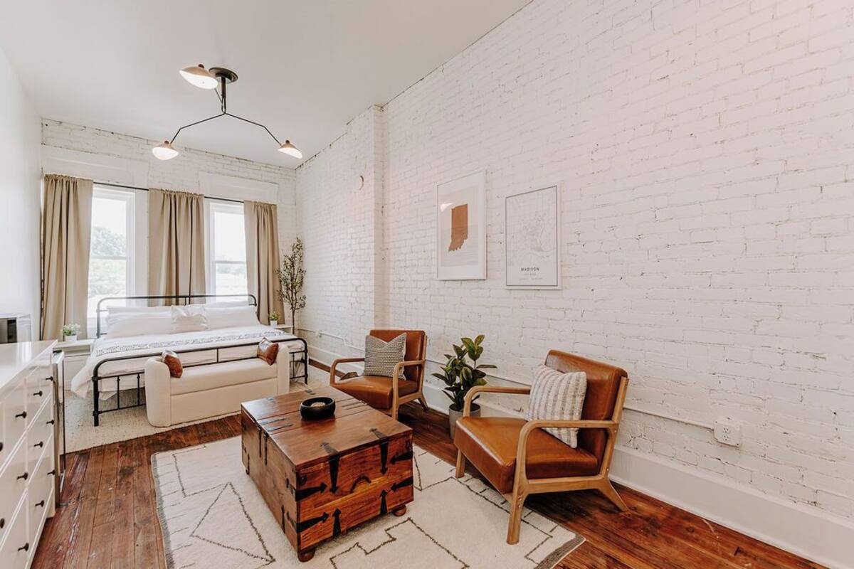 Un dormitorio moderno con pared de ladrillo blanco, cama, un cofre de madera y una zona de estar con sillas y una mesa.