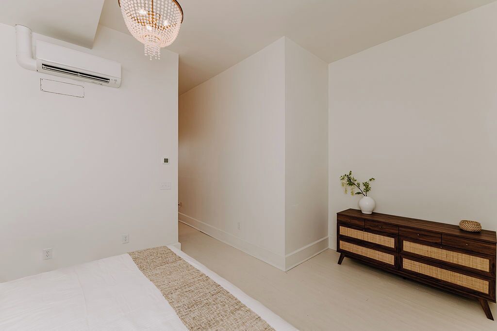 A bedroom with a bed, a wooden dresser, an air conditioning unit, and a chandelier.