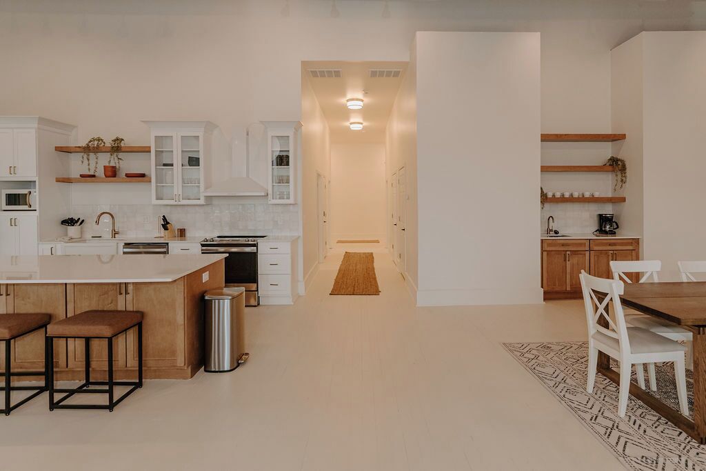 A modern kitchen with white cabinetry, a central island, and bar stools. There is a dining area with a wooden table and chairs.