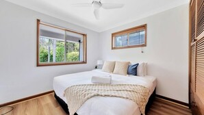 Ein Schlafzimmer mit einem Bett, einem Deckenventilator, einem Fenster mit Jalousien und Blick auf Grün.
