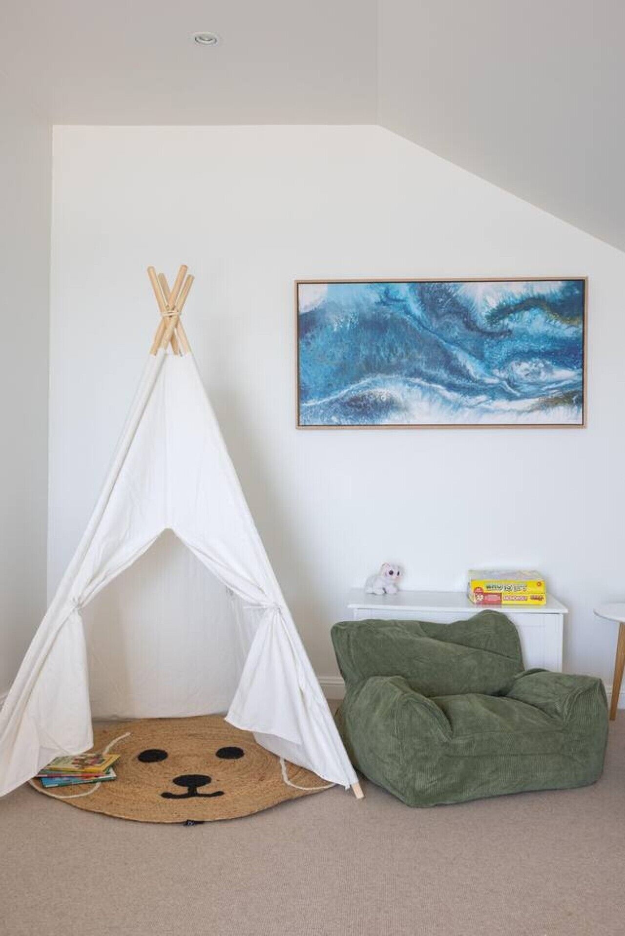 A room with a white teepee, a green bean bag, a framed abstract painting, and a small white table with a toy and a box.