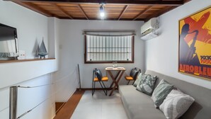 Un espacio habitable pequeño con un sofá, dos sillones, una mesita, una ventana con rejas y un equipo de aire acondicionado.