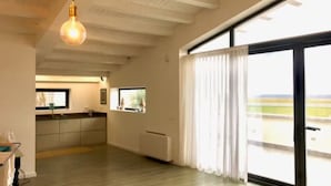A spacious room with a large sliding glass door leading to a balcony, a kitchen area with a window, and a hanging light fixture. - Mulinazzo House. Modern and stylish apartment with sea view. (Cinisi)