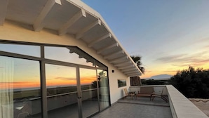 A modern balcony with glass doors, a seating area, and a view of the sunset. - Mulinazzo House. Modern and stylish apartment with sea view. (Cinisi)