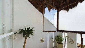 Un balcón con piso de concreto, paredes blancas y techo de paja. Hay dos plantas en macetas y paneles decorativos en la pared.