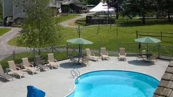 A swimming pool with lounge chairs and umbrellas, surrounded by a fenced area and houses in the background.