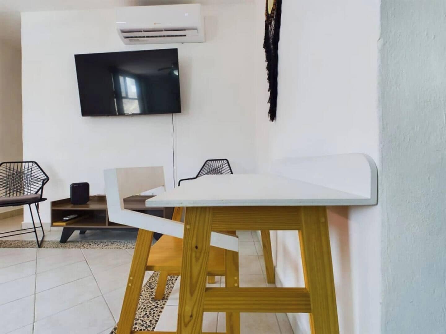 A modern living room with a white table, two black chairs, a flat-screen TV mounted on the wall, and an air conditioning unit.