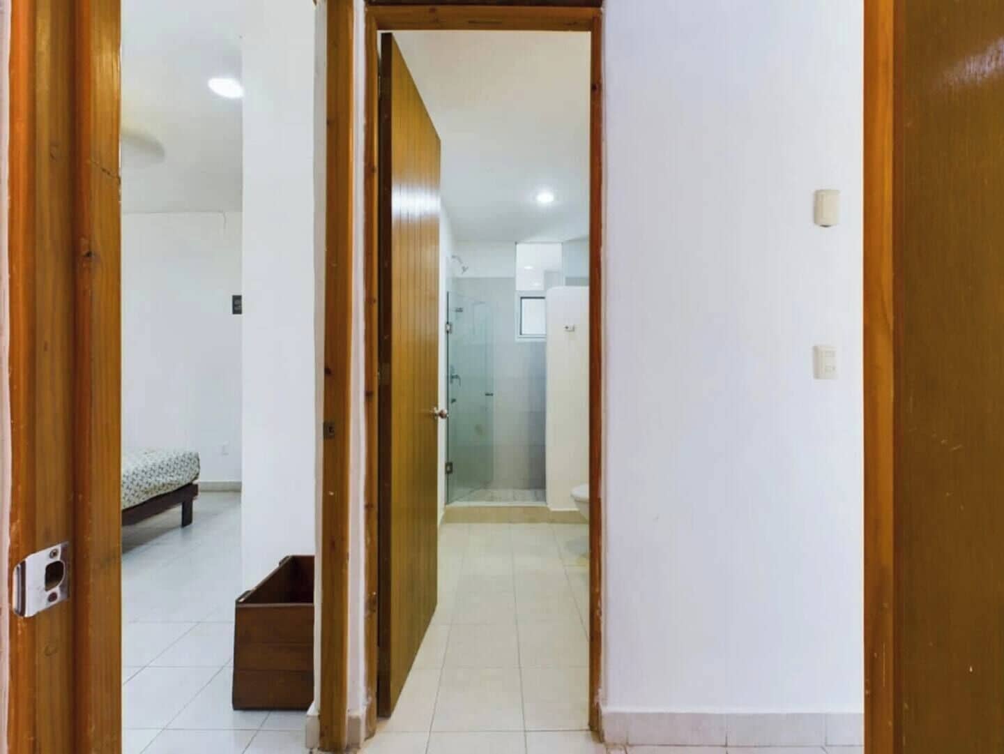 Een gang die leidt naar een badkamer met een douche en toilet. In de aangrenzende kamer staat een bed en een klein houten kastje.